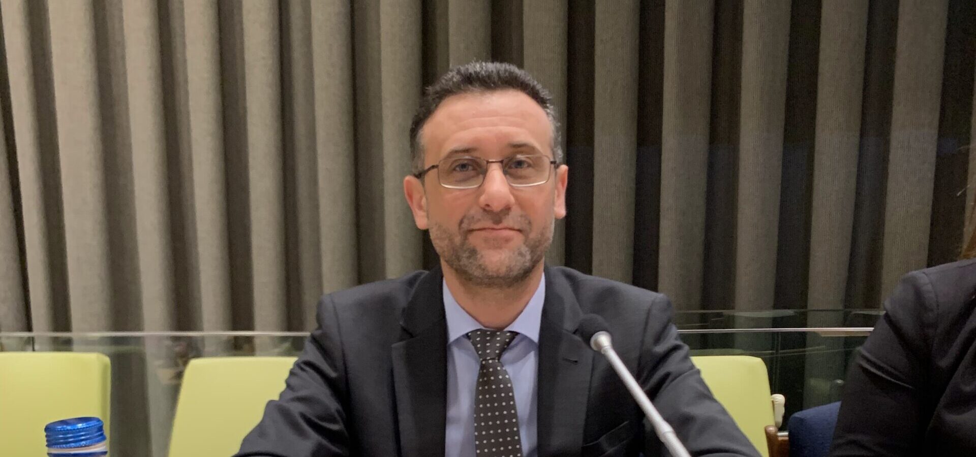 Dr Haytham Alhamwi is sitting with his arms folded on a table. He has a screen and microphone in front of him, and a sign that says Haytham Alhamwi in front of him. He is looking into the camera and smiling.