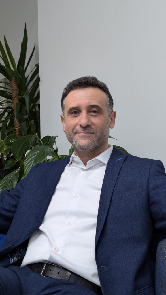 A picture of Dr. Haytham Alhamwi. He is looking directly into the camera with a small smile. He has dark hair that is greying and stubble, and is sitting on a chair in front of a white wall and plant. He is wearing a navy suit and white shirt. 