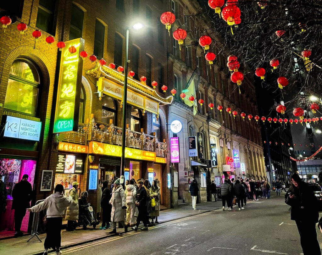 A street in Chinatown. The front of a terraced brick building is covered in signs written in Chinese characters, while red Chinese lanterns are strung overhead. Crowds of people are walking the street. It is night time. 