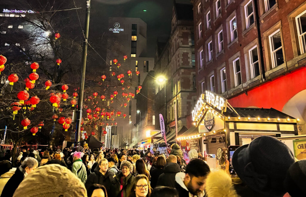 A crowd of people are swarming a street with red Chinese lanterns strung overhead. On the right of the image is the front of a terraced building with a duck wrap stall clearly visible. It is dark outside, but the street is lit with street lamps, Chinese lanterns, and food stall signs. 