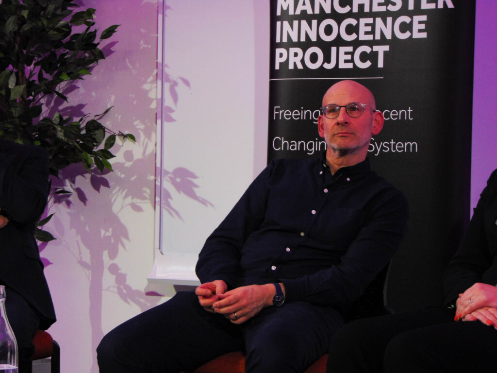 David Conn, a thin white man with a bald head, leans back on a chair with his hands on his lap. He is looking pensively above and behind the camera. Behind him is a sign that says “Manchester Innocence Project”