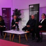 Two women and two men sit around a low table, all wearing suits. Behind them are tall sign which read “Manchester Innocence Project”. They are under stage lighting, and are looking towards an unseen crowd behind the camera.