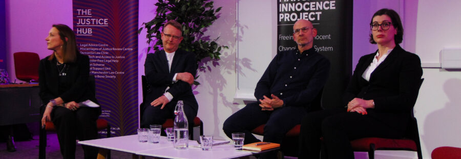 Two women and two men sit around a low table, all wearing suits. Behind them are tall sign which read “Manchester Innocence Project”. They are under stage lighting, and are looking towards an unseen crowd behind the camera.