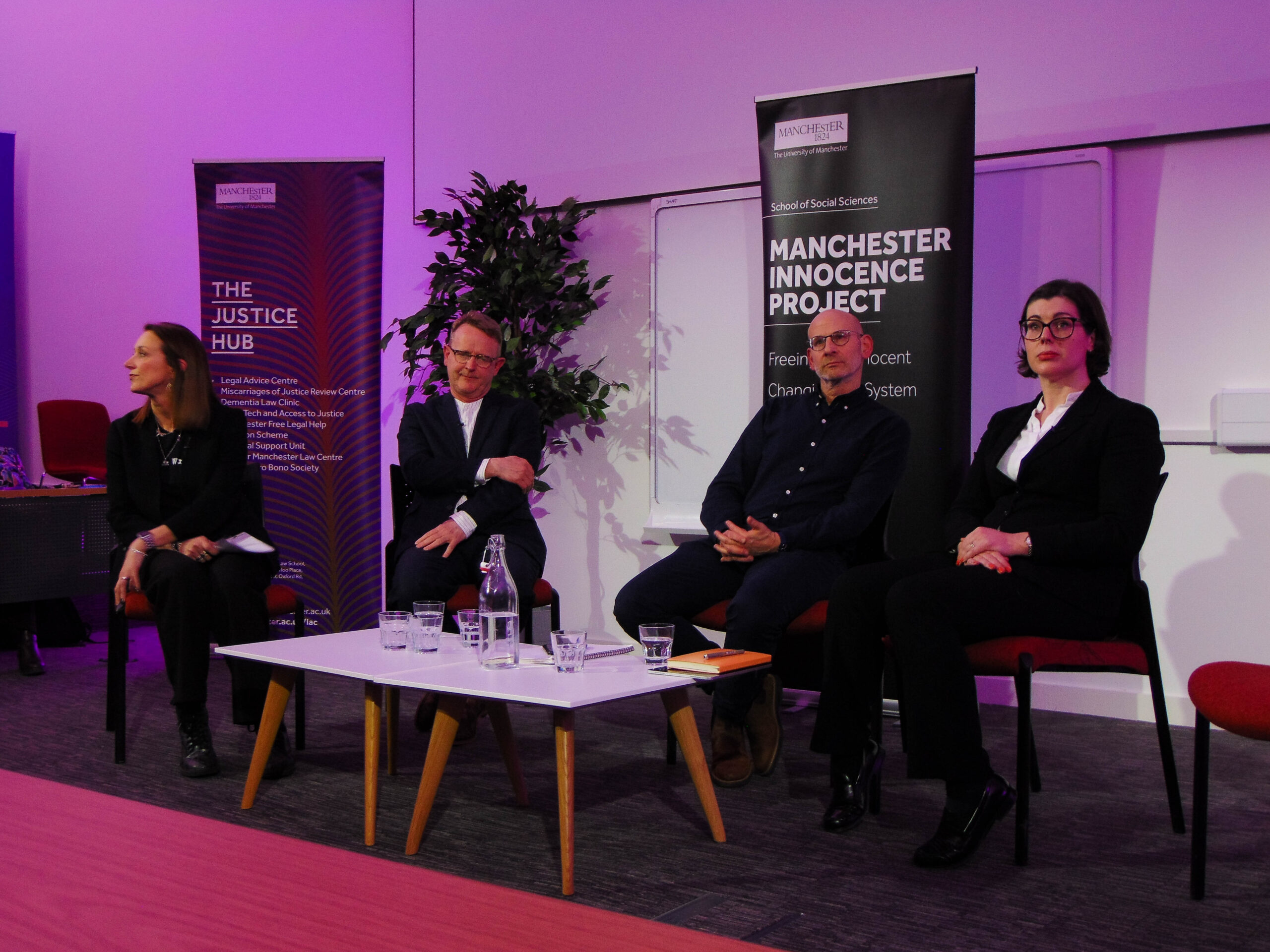 Two women and two men sit around a low table, all wearing suits. Behind them are tall sign which read “Manchester Innocence Project”. They are under stage lighting, and are looking towards an unseen crowd behind the camera.