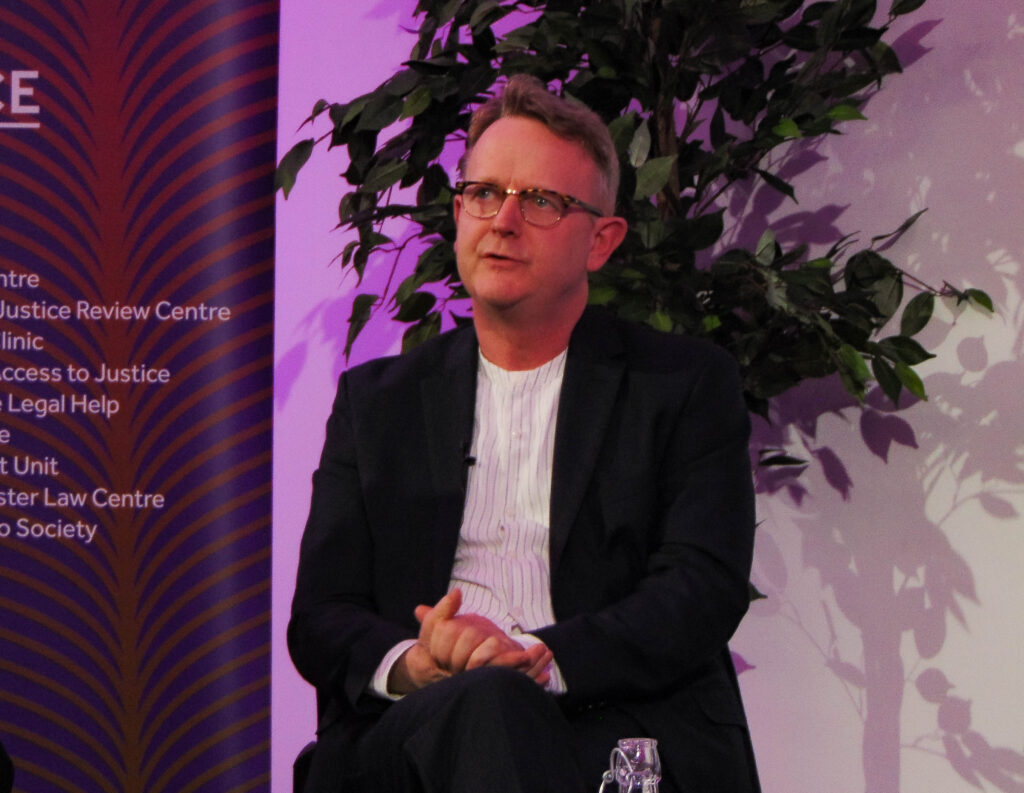 Pete Weatherby, a white man in a black blazer and white shirt, sits on a chair mid-speech with his hands clasped. 