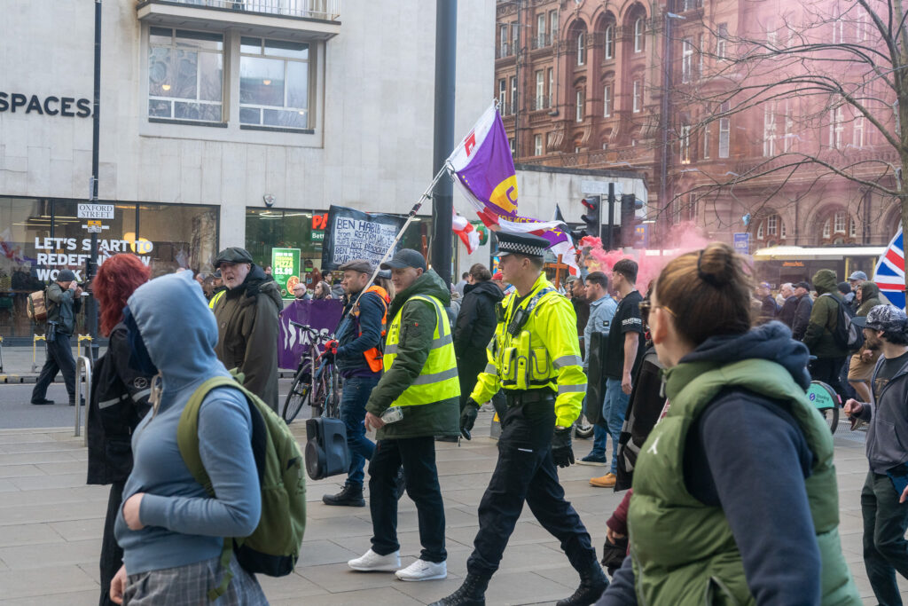 UKIP protesters walk through St. Peter's Square. In the middle, one wears a high-viz jacket and is holding a UKIP flag while being escorted by a police officer.