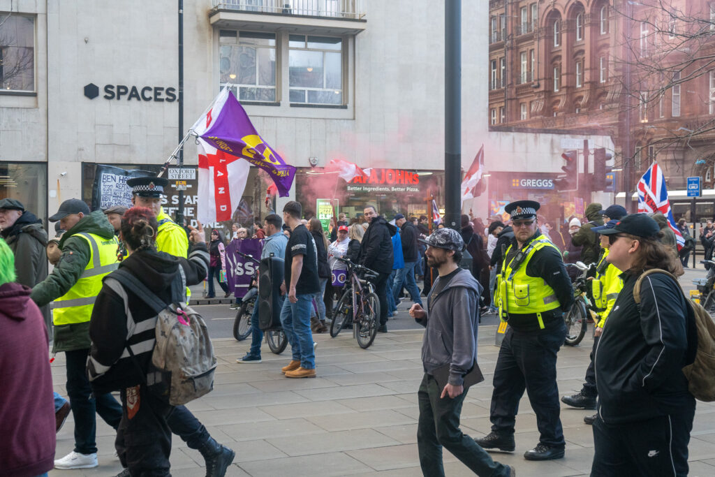 UKIP protesters walk through St. Peter's Square. In the left of the image, one wears a high-viz jacket and is holding a UKIP flag while being escorted by a police officer.