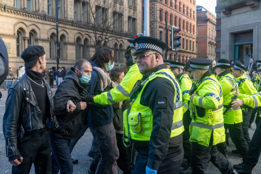 A line of police officers wearing high-vis jackets stand against a row of civilians. 