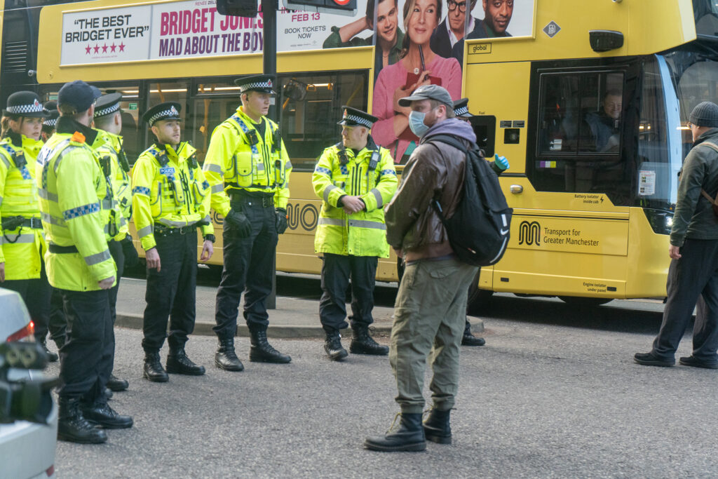 A man with a mask and rucksack stands in front of a row of officers wearing high-vis jackets.