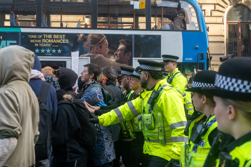 A crowd of police officers in high-vis jackets stand behind a group of counter protesters. One officer is reaching forward and touching a protester's bag.