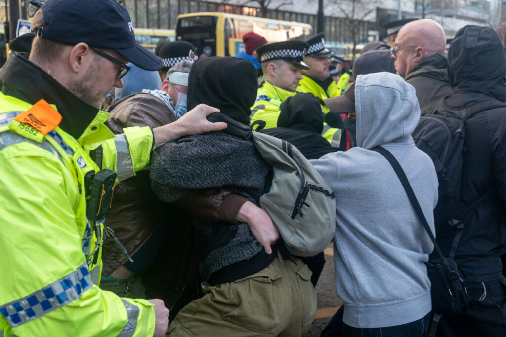 A police officer grabs a protester by the collar. They are standing in a crowd, and the protester is grabbing another protester.  