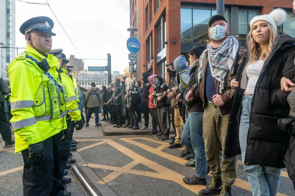 A line of police officers wearing high-vis jackets stands in front of a line of civilians. Piccadilly Gardens is visible in the background. 