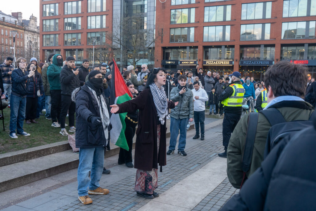 Counter-protesters stand in Piccadilly Gardens. One is shouting to the crowd and wearing a keffiyeh, while another holds a Palestinian flag. 