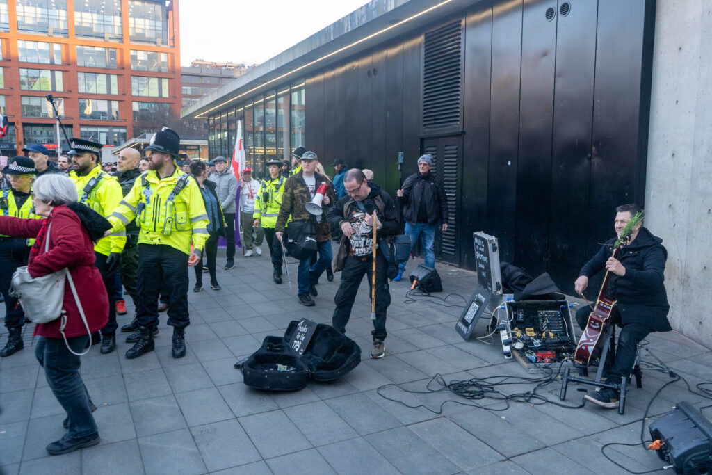 A performer sits on a stool playing a small cello and is surrounded by cables and wires. An instrument case sits in front of him. He is smiling as he looks towards the crowd of police and protesters moving past him. 