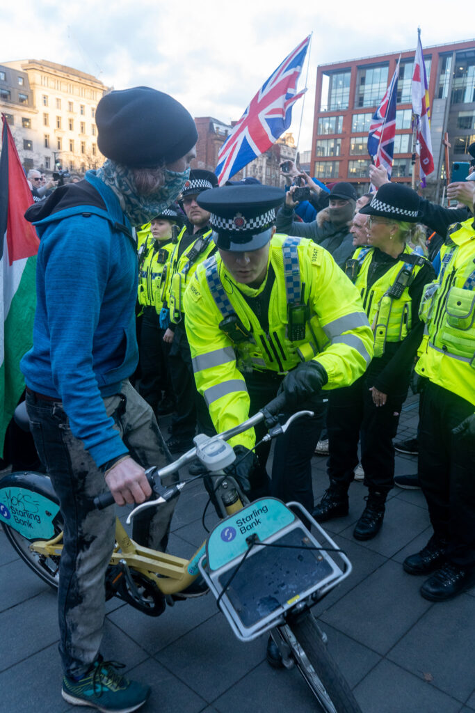 A police officer and a man both hold onto a yellow Bee bike, which the officer appears to be checking. British flags are visible in the background. 