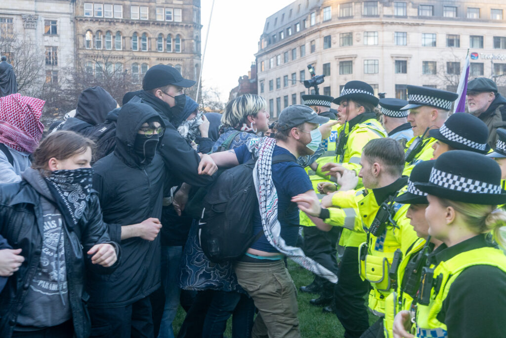 A man in a face mask and keffiyeh stands in front of a row of police officers. He appears to be moving quickly. 