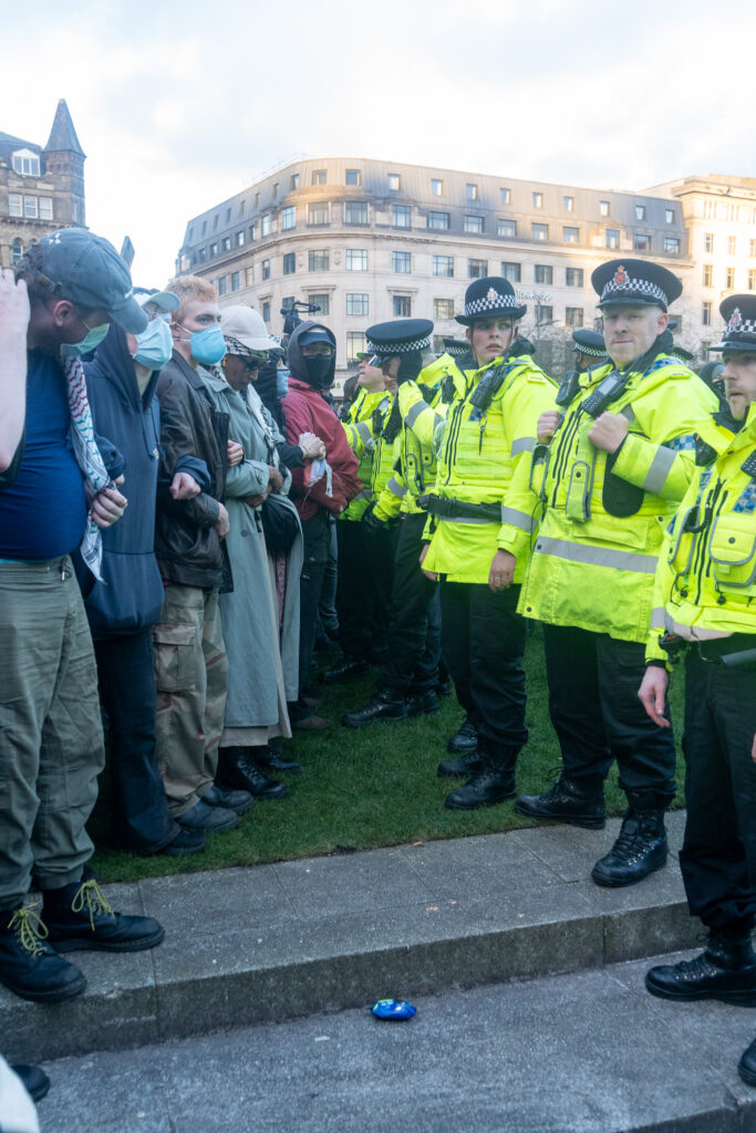 A row of police officers in high-vis jackets stand opposite a row of masked protesters in Piccadilly Gardens. 