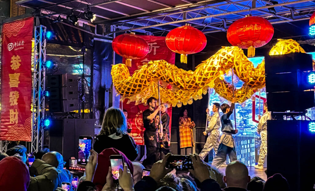 A team of people parade a golden dragon puppet above their heads on stage. There are red Chinese lanterns hanging above them, with a red Chinese banner on the left side. There is a crowd in the foreground watching the show - a little girl sits on someone's shoulders.