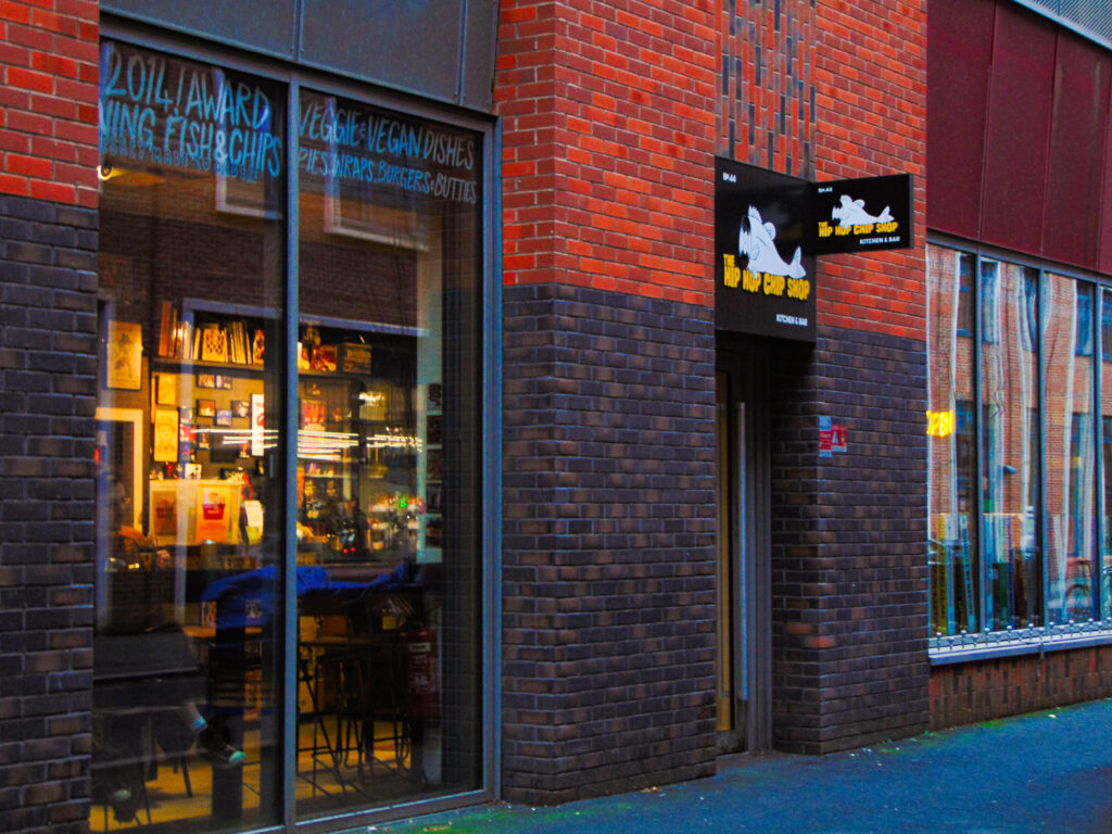 A brick shop front with a black sign reading "The Hip Hop Chip Shop" in block yellow letters. The inside of the shop is visible through a large window on the left of the image, which shows tables and a well-lit bar area filled with alcohol.