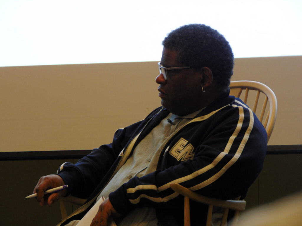 Gary Younge, a black man with short hair, glasses,, and a silver hoop earring, is facing left with a serious expression. He wears a blue jacket with white stripes, a grey button-up shirt, and is resting his arms on the arms of a wooden chair, pen in hand.