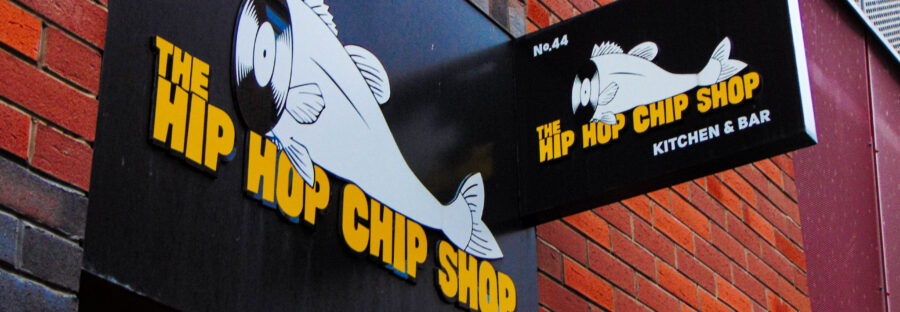 A black sign on a brick wall that reads "The Hip Hop Chip Shop (Kitchen)" in yellow block letters. The logo is a cartoon fish with the head sliced off. The neck opening is a record disk.