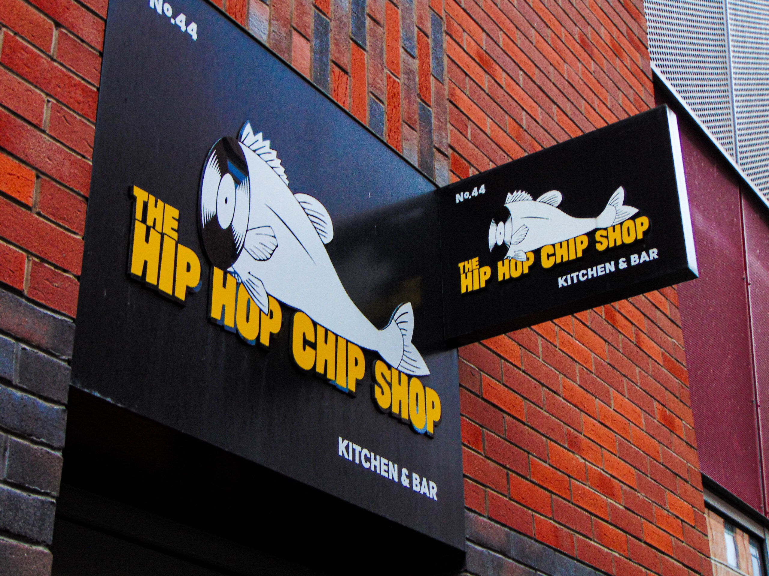 A black sign on a brick wall that reads "The Hip Hop Chip Shop (Kitchen)" in yellow block letters. The logo is a cartoon fish with the head sliced off. The neck opening is a record disk.