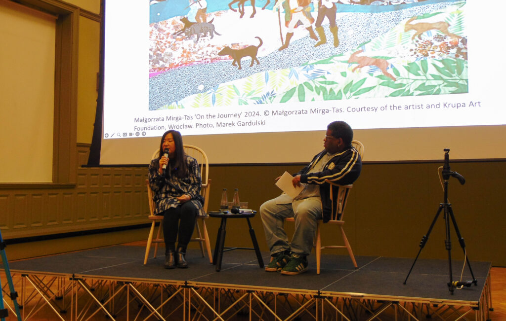 Dr. Sook-Kyung Lee, a Korean woman with long hair, a blue button-up tunic, and a microphone held to her mouth sits on a wicker chair on a stage, talking to the crowd. On the right side of the image is Gary Younge, a black man with short hair, glasses, and a blue striped hoodie and jeans. He is holding a notepad and watching Dr. Lee intently. Between them is a table with two bottles of water. Behind them is a screen with a faint art piece visible.