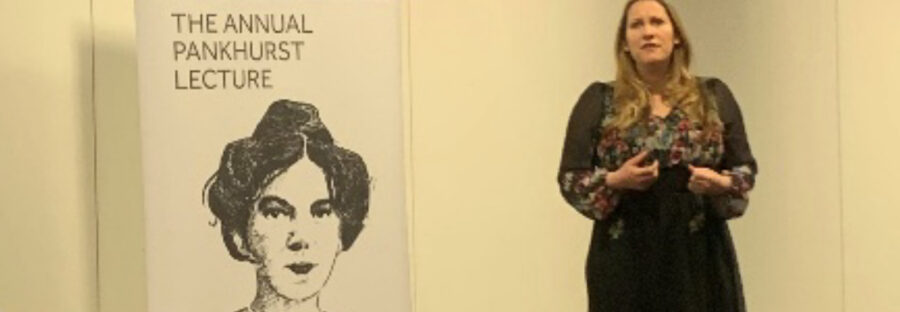 Laura Bates, a white woman with blonde hair wearing a long black dress, is mid-speech. She is standing next to a large sign on the left of the image which reads “The annual Pankhurst lecture” with a pen illustration of Emmeline Pankhurst underneath.