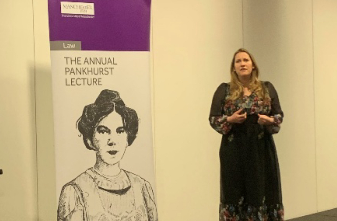 Laura Bates, a white woman with blonde hair wearing a long black dress, is mid-speech. She is standing next to a large sign on the left of the image which reads “The annual Pankhurst lecture” with a pen illustration of Emmeline Pankhurst underneath.