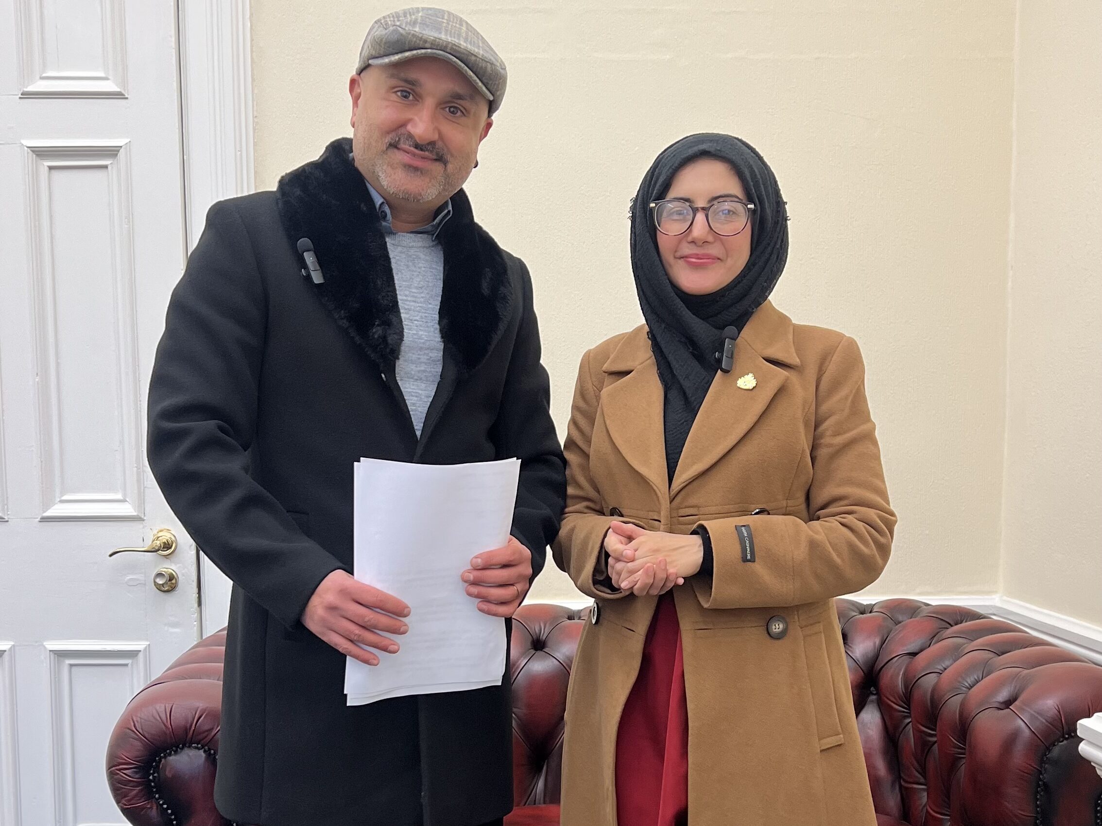 Faraz Aryan, a man in a light blue shirt, dark blue jacket, and flat cap, stands next to Cllr. Tafheen Sharif, a British-Kashmiri woman in a blue hijab and orange leather jacket. Both are smiling into the camera.