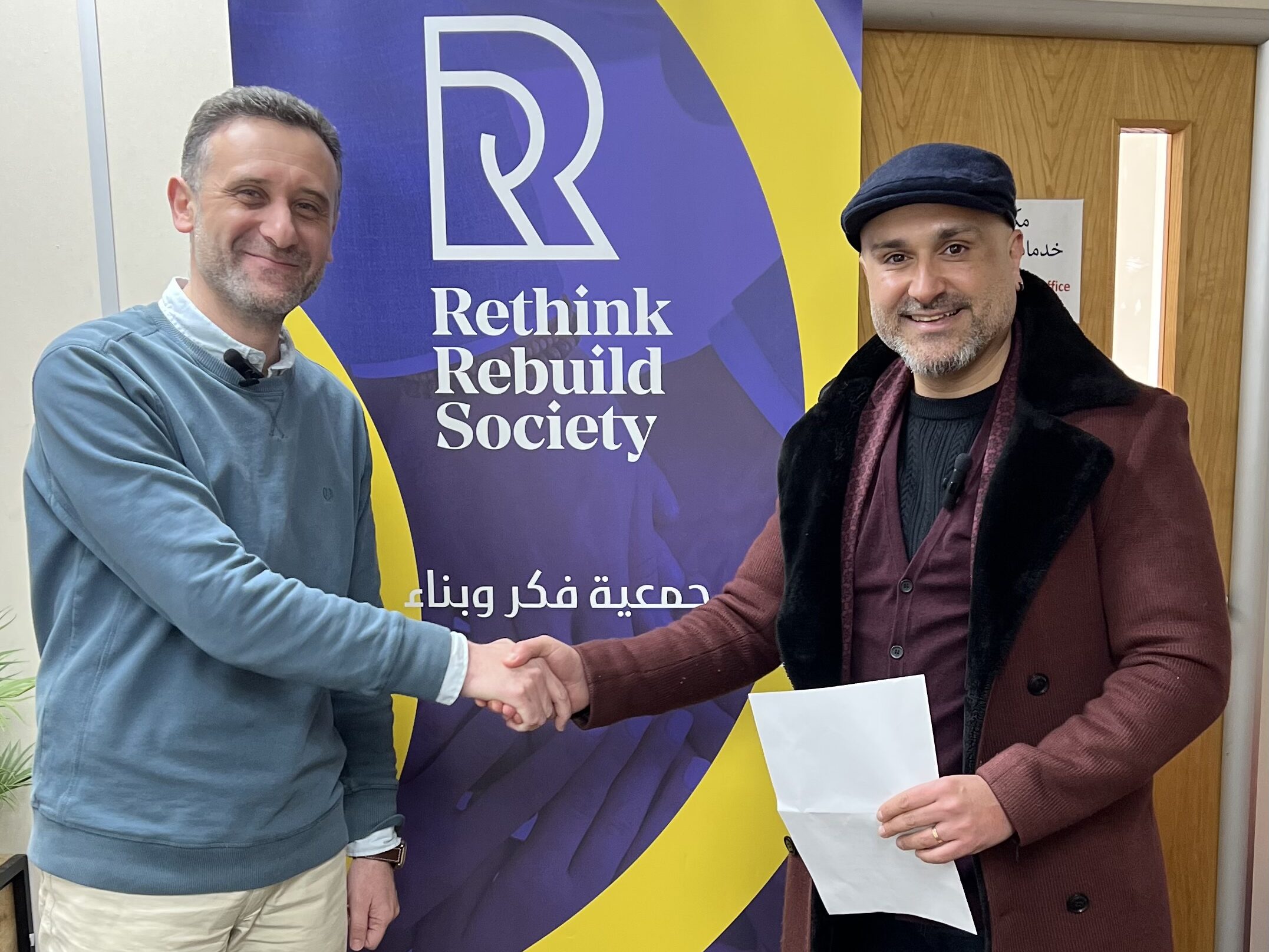 Dr Haytham Alhamwi stands on the left of the image, he is wearing a cyan jumper and cream trousers. Faraz Arian stands on the right wearing a red coat and dark suit, holding a piece of paper. The two are shaking hands in front of a banner that says Rethink Rebuild Society.