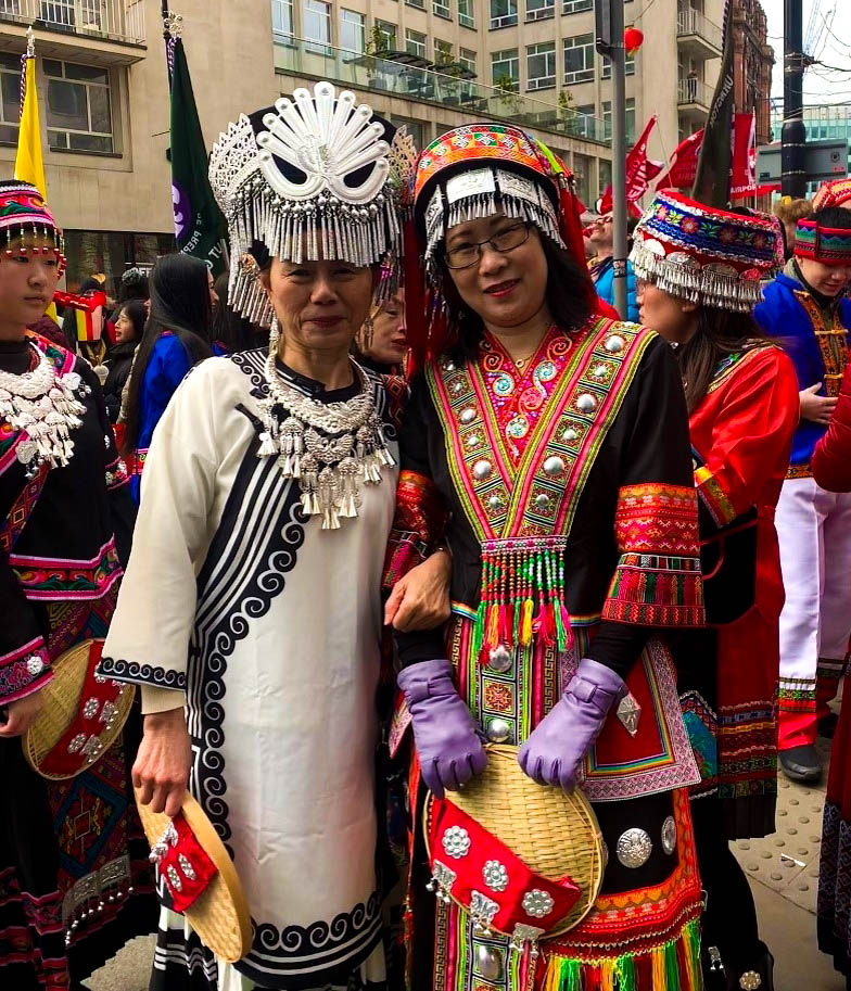 Two East-Asian women stand in front of the camera, smiling. The woman on the left is older and wears a black-and-white dress with an elaborate tasselled headpiece. She is holding the arm of the woman on the right, who wears a black-and-red dress with purple gloves. There is a crowd of people behind them wearing similar clothing.