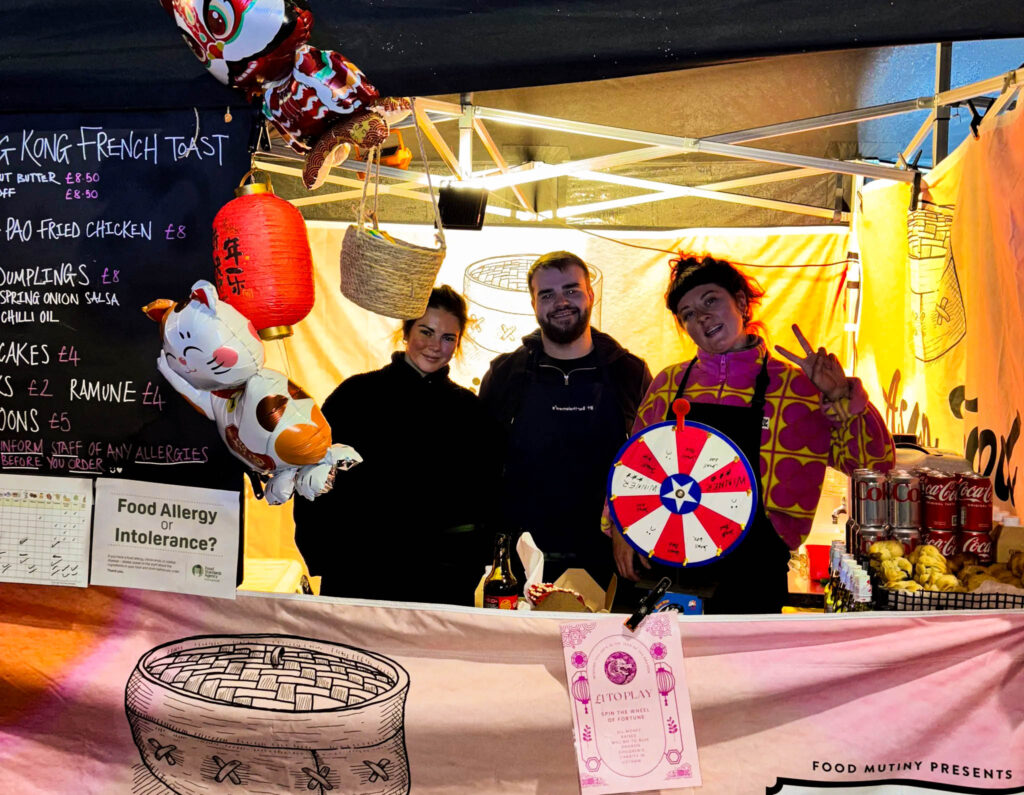 Three people stand inside a stall with a Chinese lantern, spinning wheel, and several cat balloons in front. There are two women and a man in between, all wearing black and smiling - the woman on the right also wears a pink and yellow jumper and is holding a peace sign to the camera. A blackboard on the left advertises various food dishes.