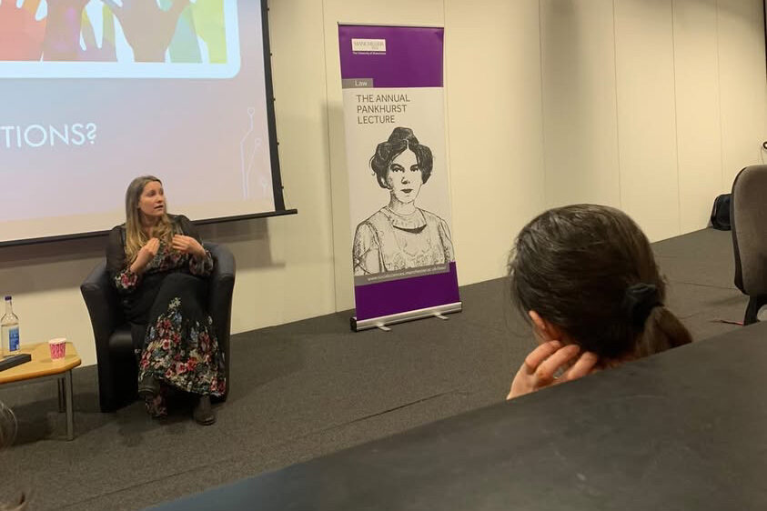 Laura Bates sits on a black chair to the left of the image, she is a white woman with blonde hair and a black floral dress. To the right is a large banner that reads 'The Annual Pankhurst Lecture' with an illustration of Emmeline Pankhurst. 