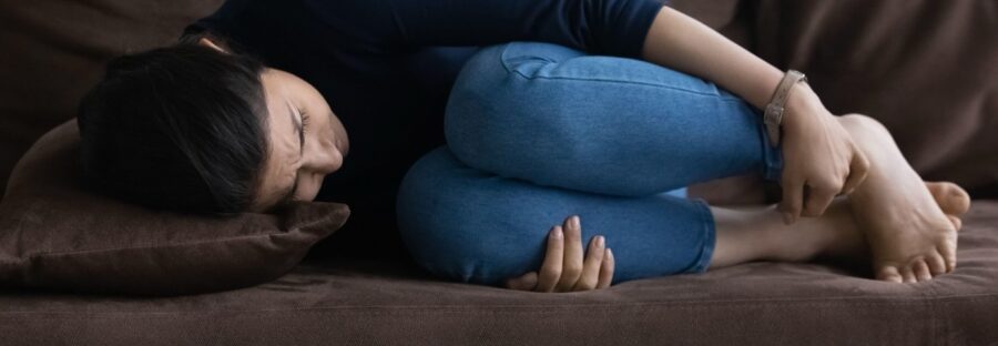 A woman in a dark shirt and jeans lies on her side on a sofa, clutching her knees in a foetal position.