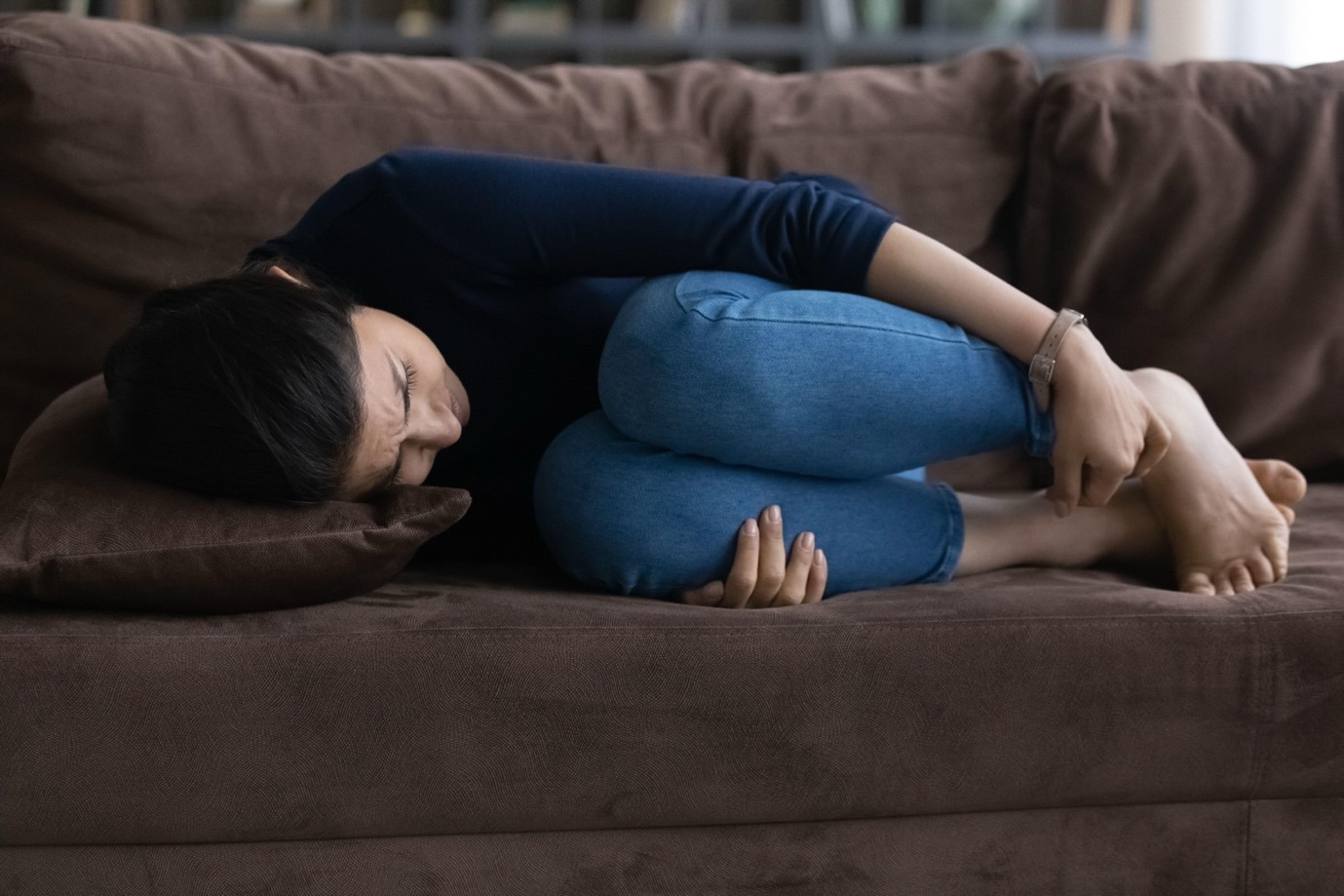 A woman in a dark shirt and jeans lies on her side on a sofa, clutching her knees in a foetal position.