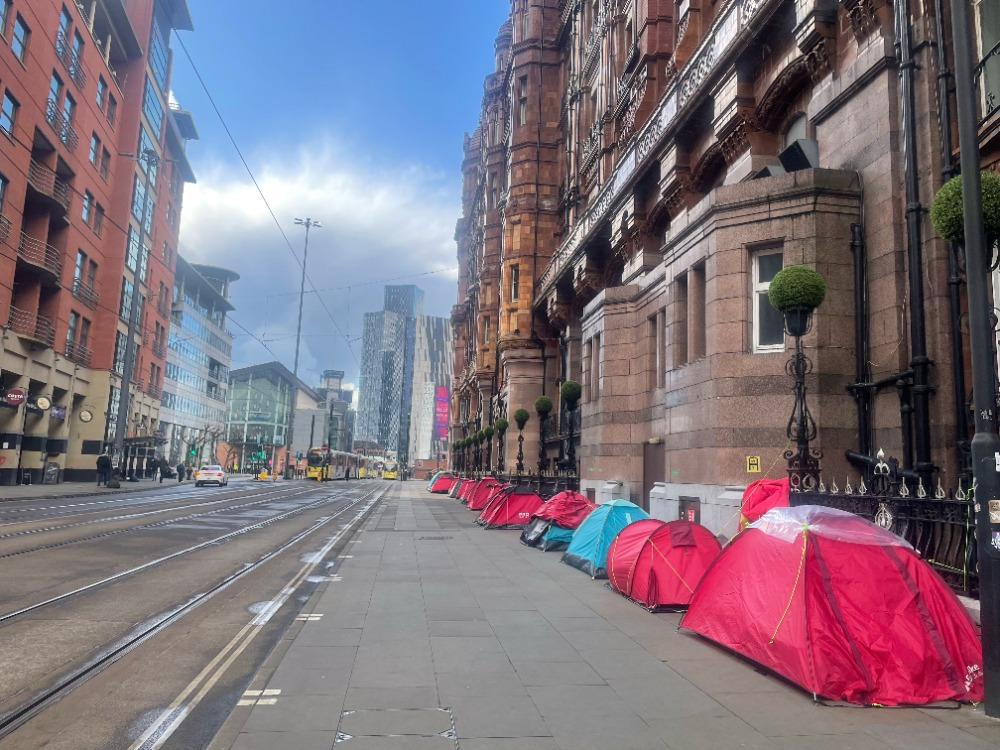 A row of tents lining the road leading out of St. Peter’s Square. The tents stand next to a large Victorian brick hotel, while skyscrapers are visible in the background. There are no people in the image.