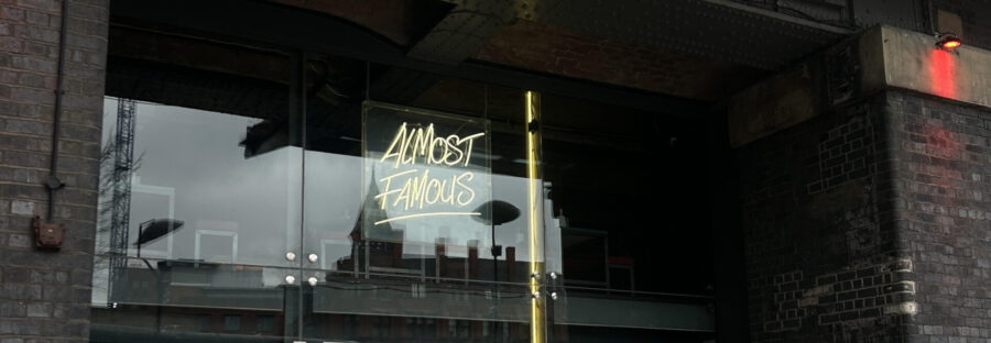 The doorway of the Almost-Famous burger restaurant. It is dark inside with a glass door and window, with the inside not visible. A glowing yellow neon sign reads Almost Famous.