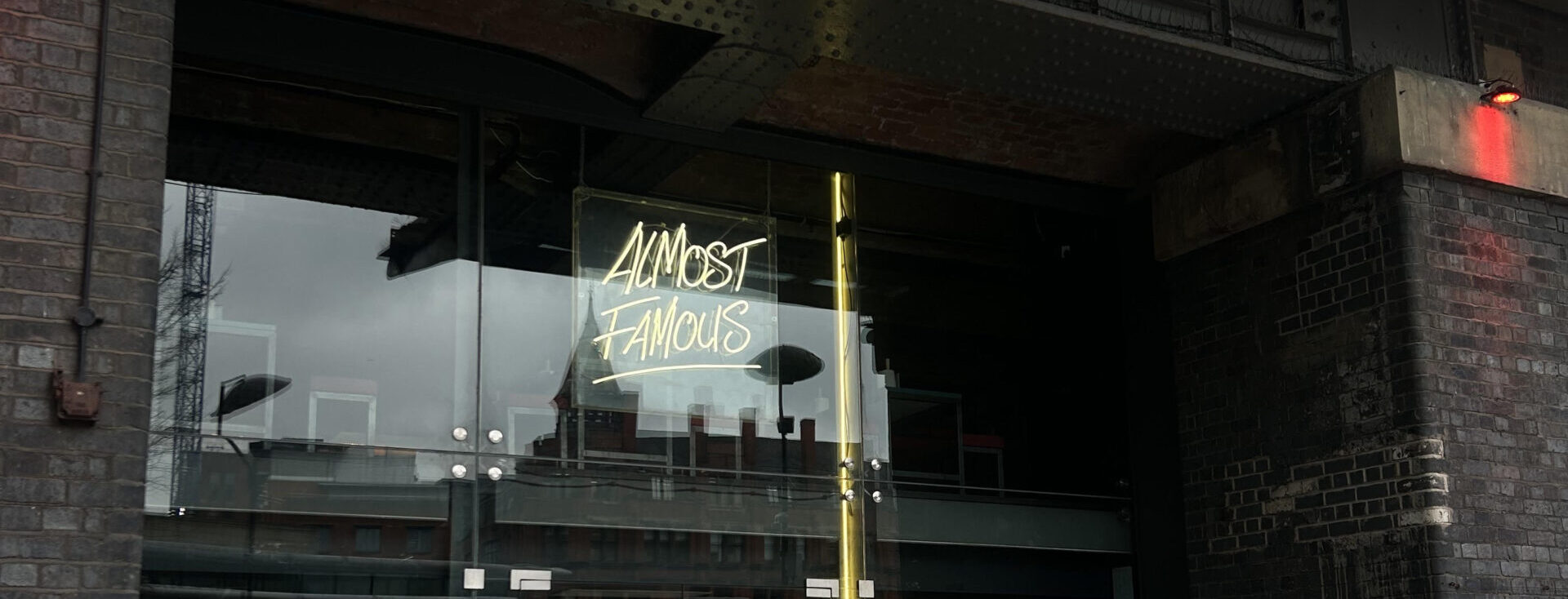 The doorway of the Almost-Famous burger restaurant. It is dark inside with a glass door and window, with the inside not visible. A glowing yellow neon sign reads Almost Famous.