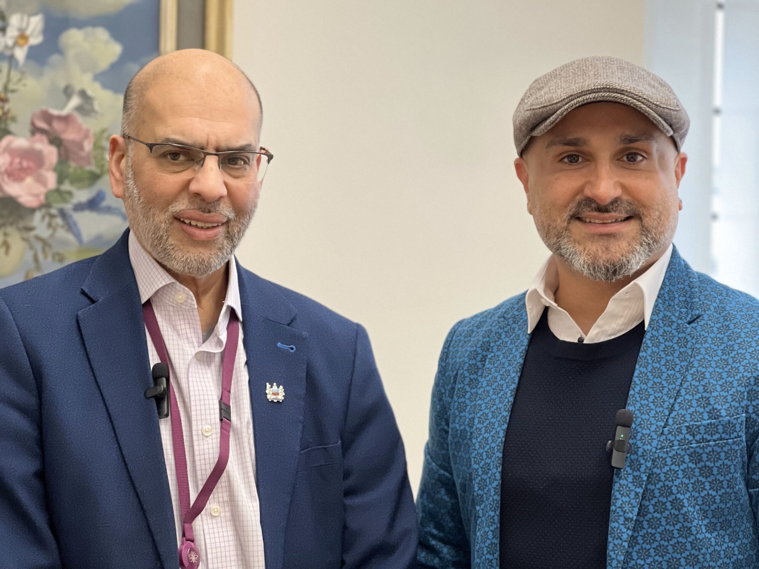 Rabnawaz Akbar stands on the left of the image. He is bald and wears glasses, and is smiling at the camera. He is wearing a pink shirt, blue suit, and a lanyard. Faraz Arian stands to the right of the image, wearing a blue suit, dark jumper, white shirt, and grey flatcap. He is holding a sheet of paper and smiling at the camera.