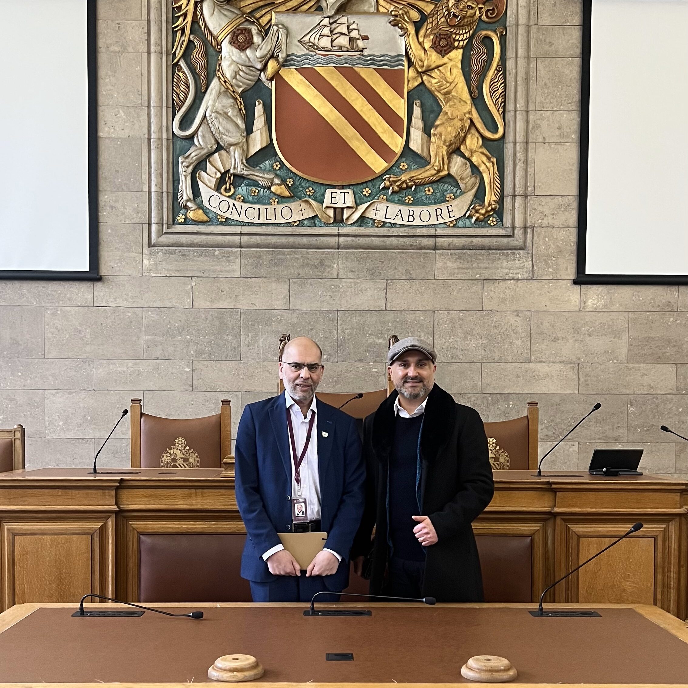 Rabnawaz Akbar stands on the left of the image. He is bald and wears glasses, and is smiling at the camera. He is wearing a pink shirt, blue suit, and a lanyard. Faraz Arian stands to the right of the image, wearing a blue suit, dark jumper, white shirt, and grey flatcap. He is smiling at the camera. they are stood in a council chamber with grey brick walls, oakwood tables, and a medieval sign on the wall behind. The sign reads: Concilio et labore'