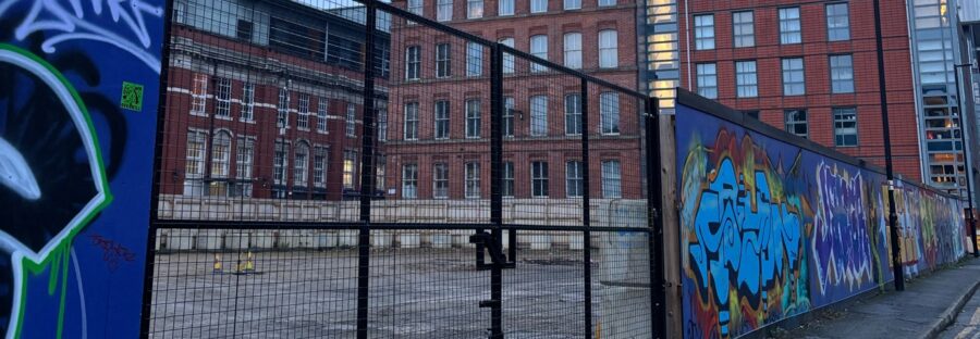 A metal gate leading into an empty concrete area. Tall, red-brick buildings are visible in the background. The gate is set into a brick wall that leads further into the distance, covered in graffiti.