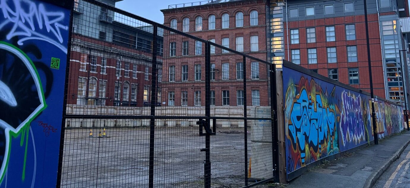 A metal gate leading into an empty concrete area. Tall, red-brick buildings are visible in the background. The gate is set into a brick wall that leads further into the distance, covered in graffiti.