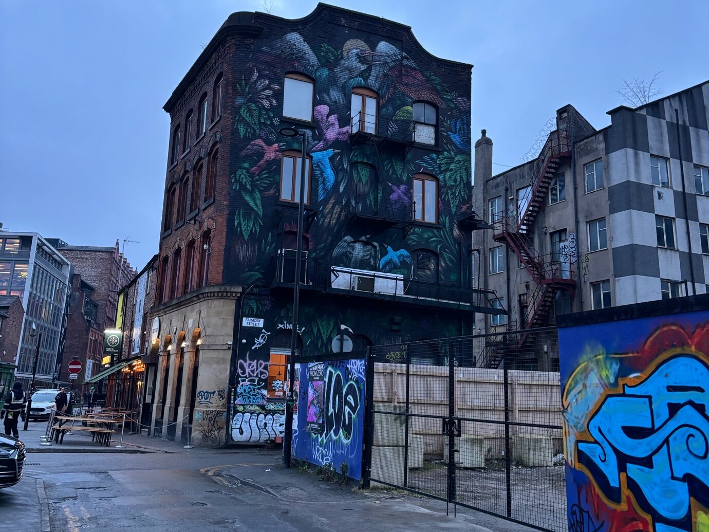 A metal gate leading onto an empty concrete area. It is set into a wall covered in graffiti. A tall building overlooks the area, also covered in graffiti. The pavement leads into the distance, with lantern-lit shops and wooden tables visible in the distance.