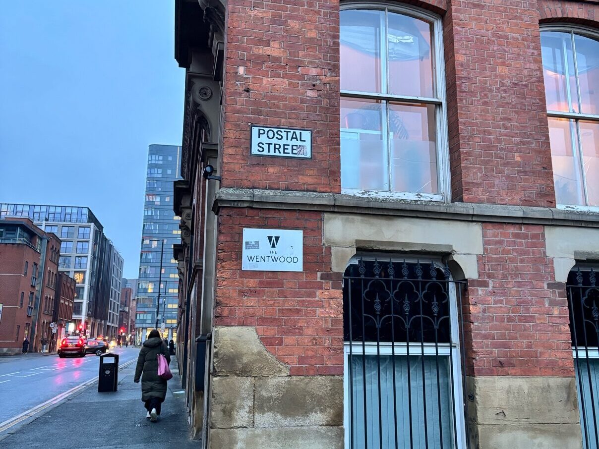 A red brick building n the corner of the street. It has a street sign that says ‘Postal Street’, above another sign that says ‘The Wenworth’. Skyscrapers are visible in the background down the road.