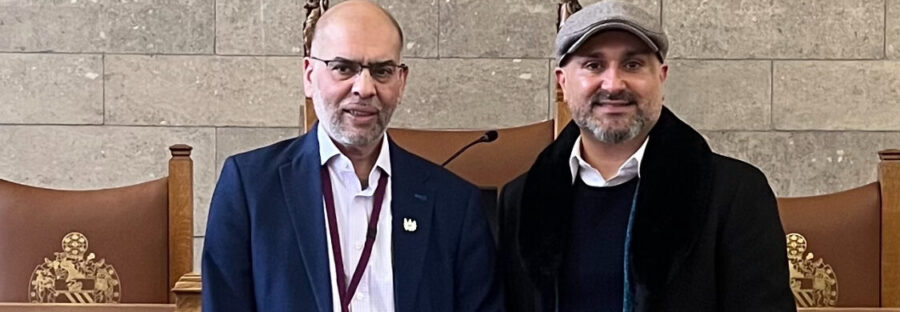 Rabnawaz Akbar stands in a navy blue suit and white shirt next to Faraz Arian, who wears a grey hat and dark coat. they are standing in front of oak panelled pews and a grey brick wall.