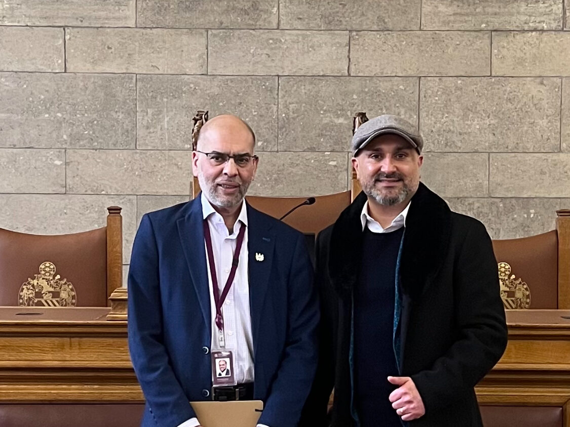 Rabnawaz Akbar stands in a navy blue suit and white shirt next to Faraz Arian, who wears a grey hat and dark coat. they are standing in front of oak panelled pews and a grey brick wall.