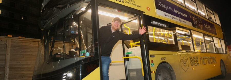 Ian Burke wears a black jacket and jeans as he waves out of the doors of a yellow Bee Network bus at night.