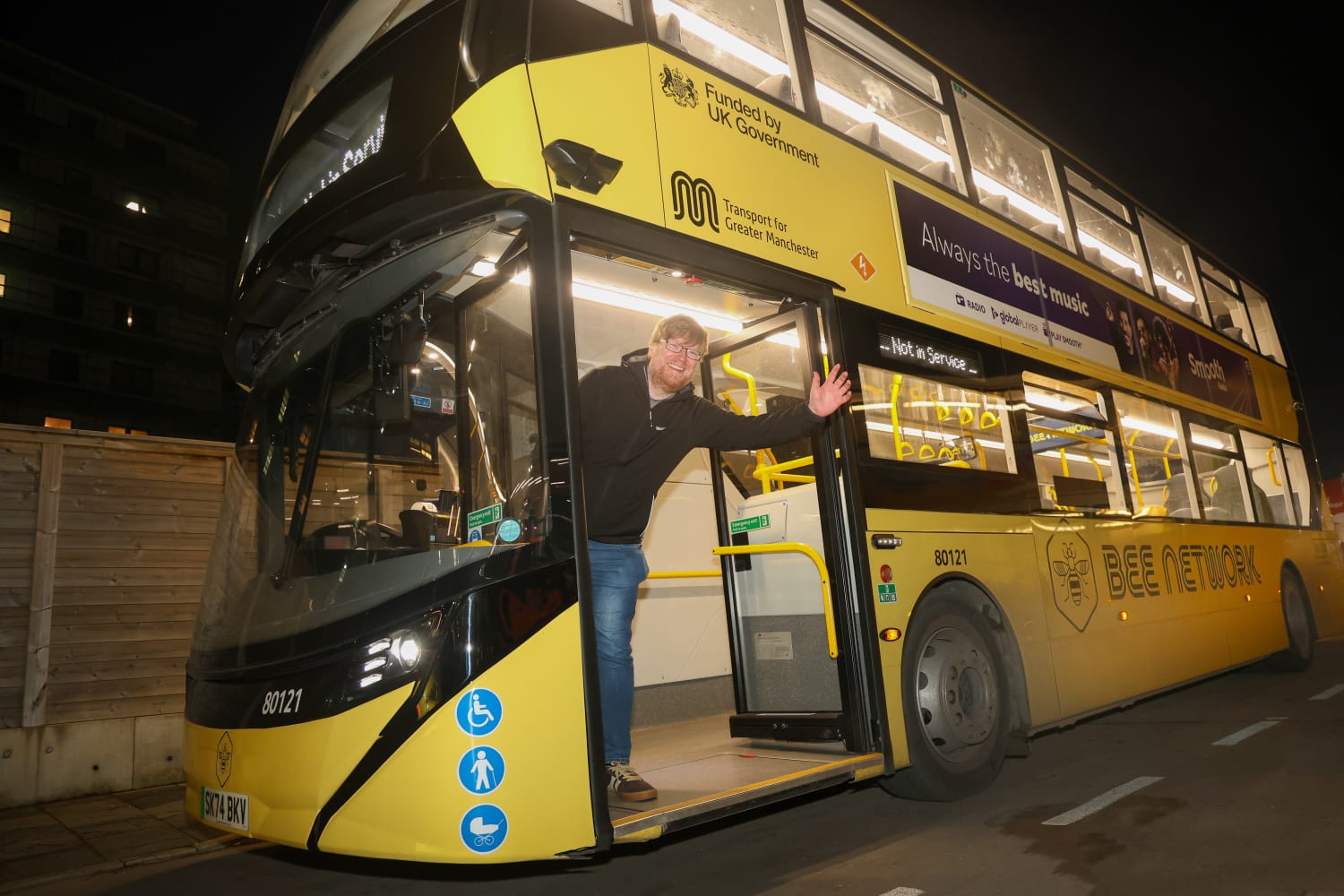 Ian Burke wears a black jacket and jeans as he waves out of the doors of a yellow Bee Network bus at night.