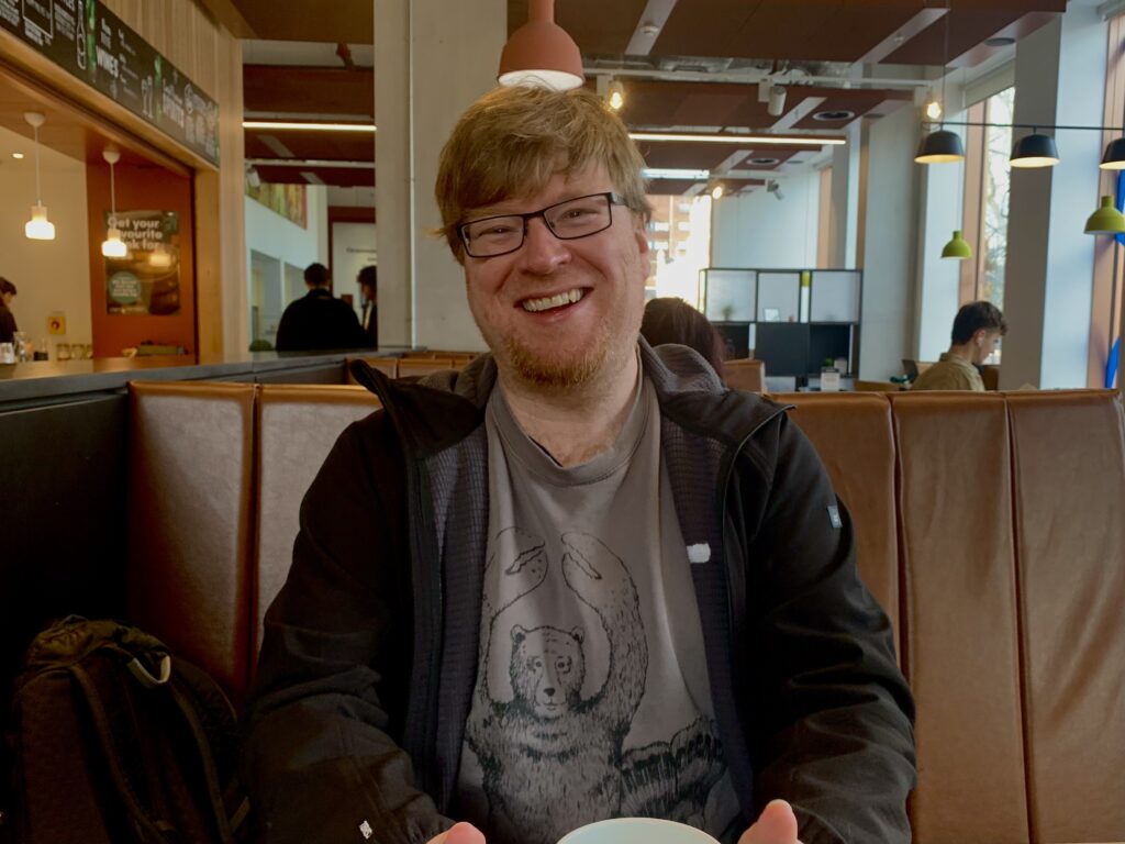 Ian Burke, a man with ginger hair, glasses, and a grey shirt, smiles into the camera. He is sat in a warmly-lit café.